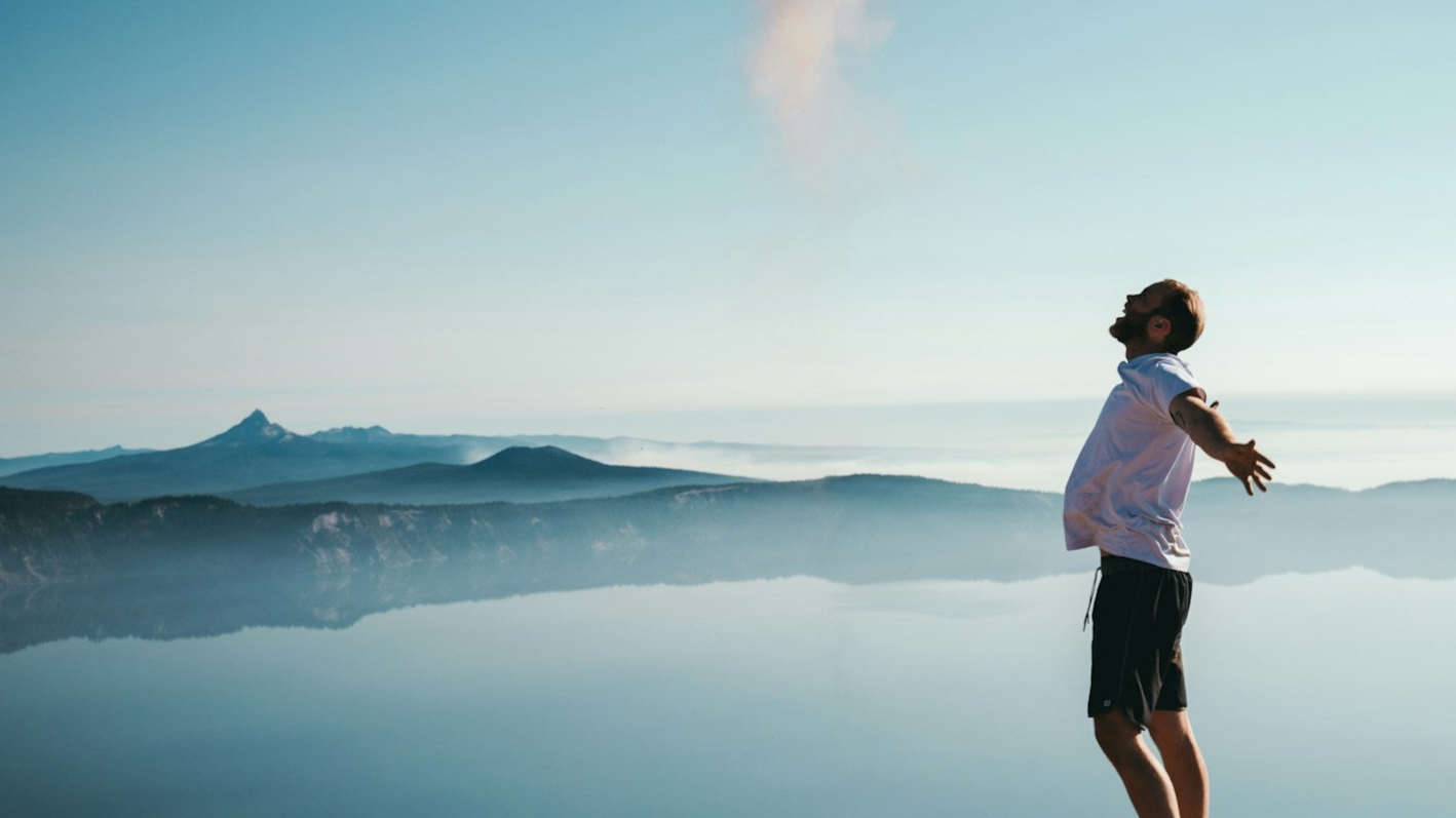 A person in a white shirt and black shorts standing with their arms stretched on a mountain top, overlooking a lake and mountain range.